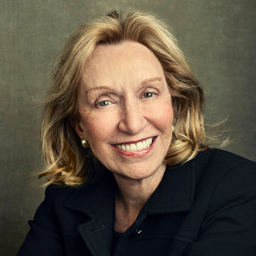 A smiling portrait of Dris Kearns Goodwin with an olive green background. Doris is a white woman with dirty blonde mid-length hair. She wears pearl earrings and a black trench coat.