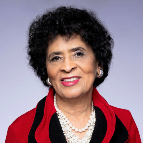 A smiling portrait of Dr. Thelma Daley sits on a light purple gradient background. Thelma is a Black woman with very curly and short black hair. She wears pink-red lipstick with multiple strings of pearls and chunky silver hoop earrings. Her blazer is black and red.