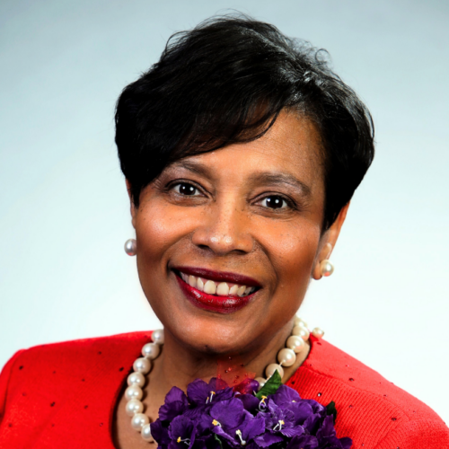 A smiling portrait of Elsdie Cooke-Holmes sits on a pale blue gradient background. Elsie is a Black woman with a short, Black pixie cut. She wears big pearl earrings and a chunky pearl necklace. Her lipstick is maroon. She wears a bright red blazer adorned with a bouquet of purple flowers.