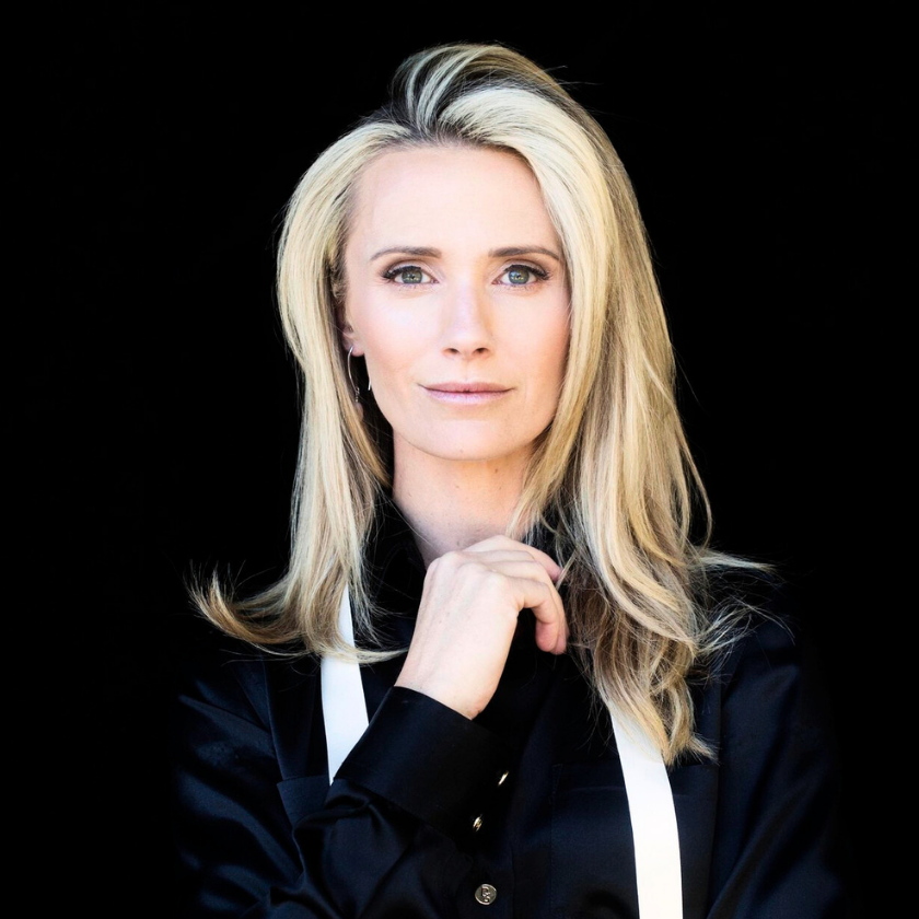 A serious-faced portrait of Jennifer Siebel Newsom on a black background. She has her hand poised near her chin. She stares directly in the camera and is a white woman with silver blonde hair with dark streaks. She wears a black button-down with white suspenders.