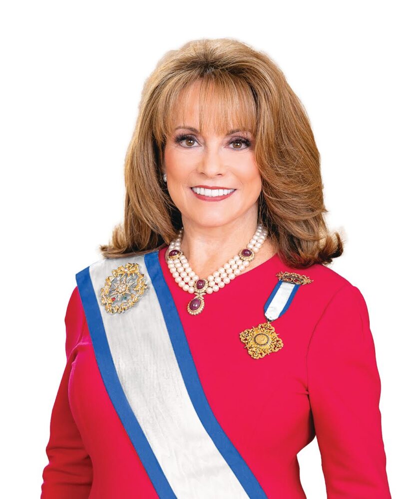A smiling portrait of Pamela Wright who is a white woman with mid-length voluminous brown hair. She wears a bright red dress adorned with blue and white-striped sashes and gold emblems. She has a necklace with red jewels and chunky pearls.