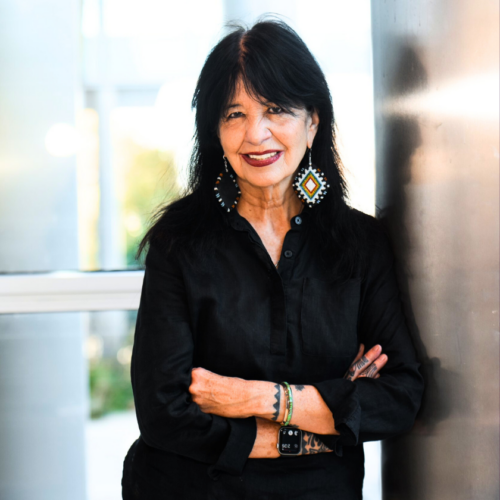 Joy Harjo leans against a wall with her arms crossed and smiles. She wears colorful, beaded dangling earrings and rainbow bracelets adorn her wrists. She wear a black button-down and wears her dark black hair down.