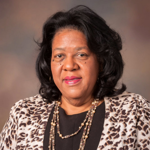 A close-mouth smiling portrait of Karen Hill. Karen wears a cheeta-print cardigan over a black shirt and gold, beaded necklace. She is a black woman with curly, mid-length black hair.