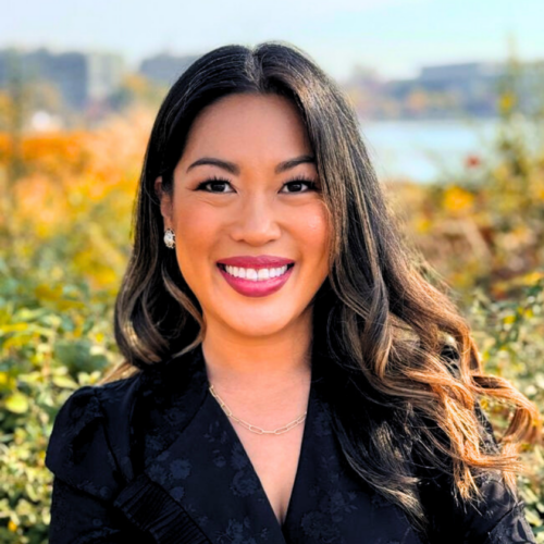 A smiling portrait of Kay Durante, an Asian woman with long, black and curled hair, who wears a black blazer and appears to be in front of a river and natural foliage.