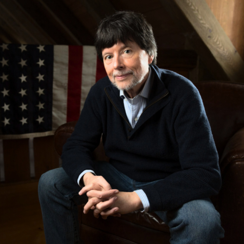 Ken Burns sits on an armchair with his hands out in front of him. He has a closed mouth smile, dark short hair, and a slight beard. He wears a striped button down with dark pullover over top. He sits in front of an American flag.