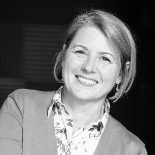 A black and white smiling portrait of Kerri Hoffman. Hoffman is a white woman with a short bob cut. She wears a floral, patterned button-down and plain blazer.