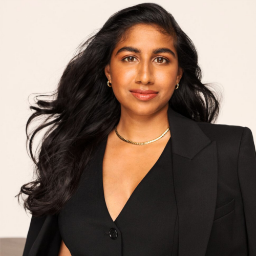 Monica Padman has a closed mouth smile and stands with her arms crossed in a powerful pose. She is an Indian women with long black hair that blows, majestically. She wears a black vest and black blazer with a simple, gold chain necklace.