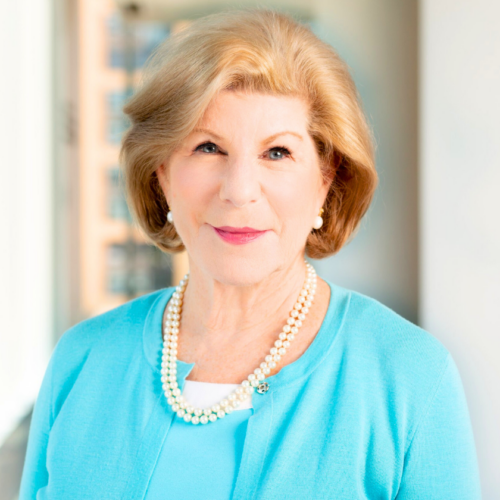 A portrait of Nina Totenberg with a closed-mouth smile. She is a white woman with honey blonde hair in a fluffy bob. She appears to stand in front of a window. She wears pearl earrings and a layered pearl necklace. Her cardigan is bright teal with a matching shirt.