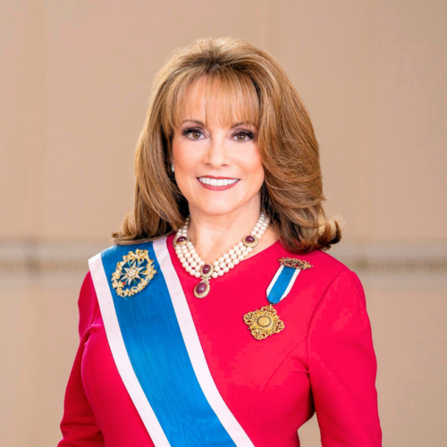 A smiling portrait of Pamela Wright who is a white woman with mid-length voluminous brown hair. She wears a bright red dress adorned with blue and white-striped sashes and gold emblems. She has a necklace with red jewels and chunky pearls.