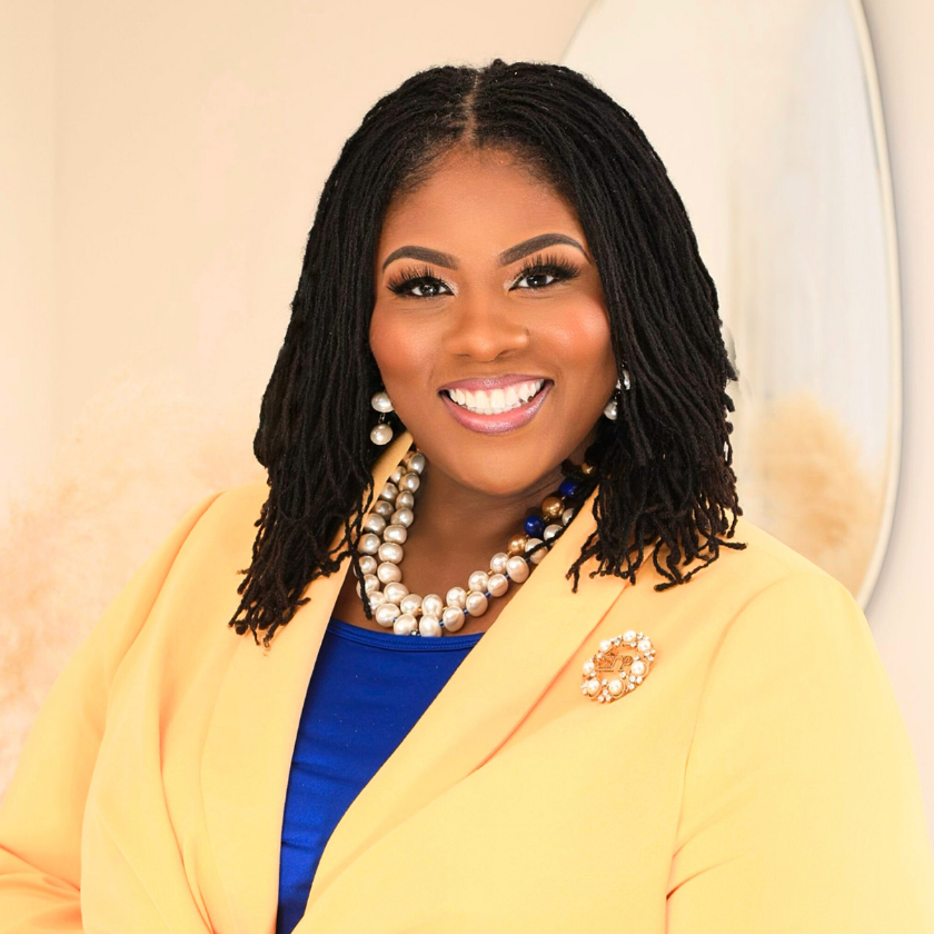 Rasheeda S. Liberty smiles in front of an oval mirror. She is a Black woman with very curly, mid-length hair. She wears a chunky pearl necklace, bright blue shirt, and creamy yellow blazer over top.