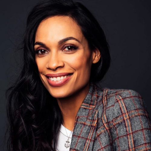 A smiling portrait of Rosario Dawson sits on a dark gray background. She has long and slightly curled Black hair which is behind one ear and falling over her left shoulder. She wears a white t-shirt under a plaid blazer.