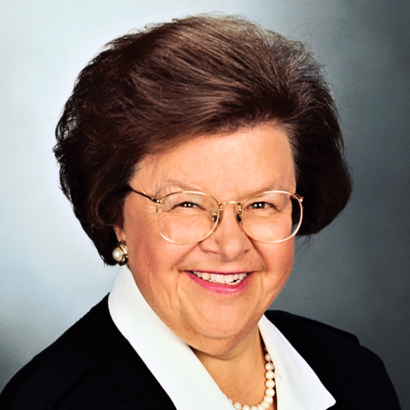 A portrait of Senator Barbara Mikulski sits on a gray, gradient background. She smiles and has big, wire-frame square glasses. Her hair is short and voluminous. She wears pearl earrings and a string of pearls around her neck. She wears a white button-down and a black blazer.