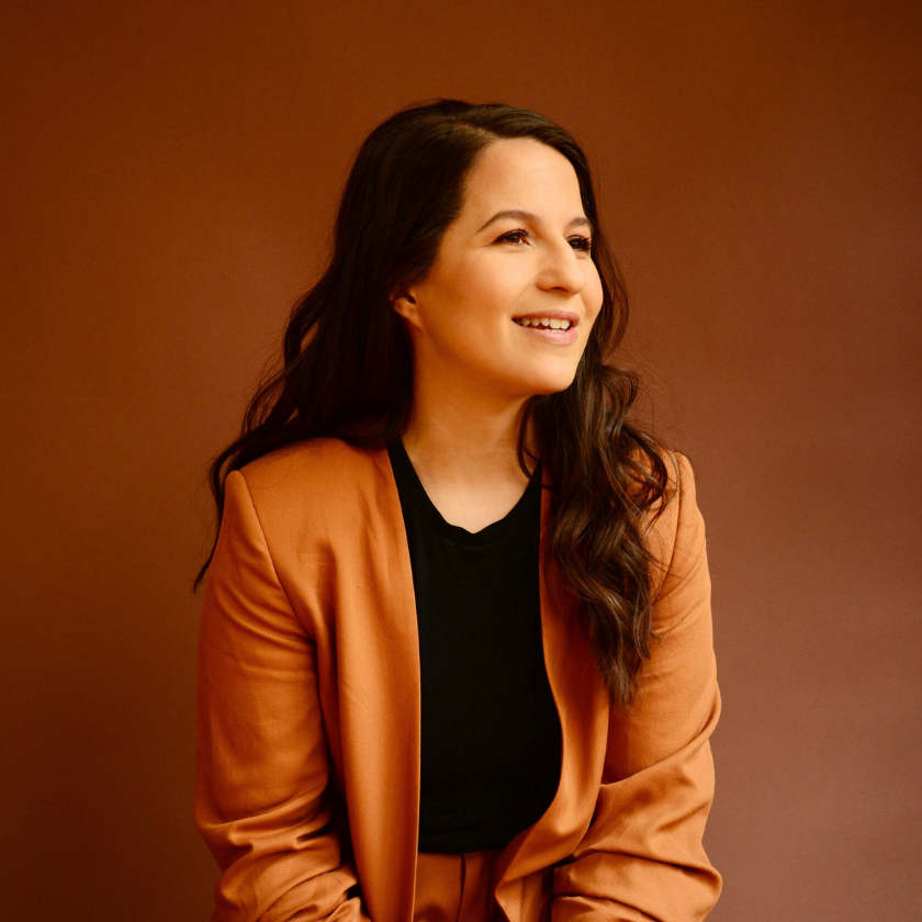 A portrait of Shaina Taub smiling and looking off to the right is on a brown ombre background. She has brown curly hair and wears a camel-colored suit with a black shirt.