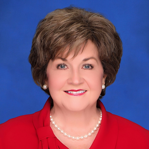 A smiling portrait of Suellen Brazil, a woman with puffy brown hair and who wears pearl earrings, a string of pearls, and bright red blazer sits on a blue background.