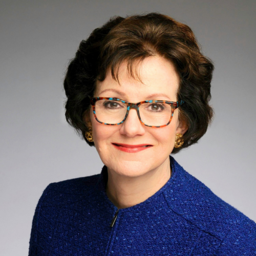 A closed mouth smiling portrait of Susan Combs sits on a gray, gradient background. She wears tortoise-shell glasses and coral lipstick. Her brown hair is styled in a short and curled brown bob. She wears a textured, blue zipped blazer.