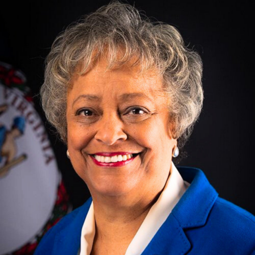 A smiling portrait of the Honorable Kay Cole James sits on a background with the Virginia flag. She wears red lipstick and has short, gray hear. She wears a bright blue blazer and white top.